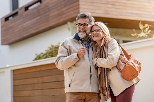 Senior couple embracing and smiling in front of a modern house on a sunny day, showcasing warmth, love, and happiness in a relaxed outdoor setting.