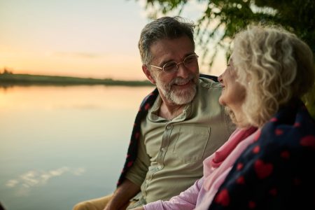 Affectionate senior couple is enjoying a peaceful evening by the lake, wrapped in a blanket and sharing a tender moment as the sun sets