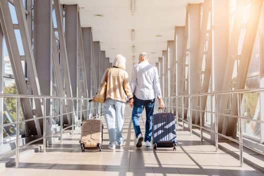 Happy mature couple walking hand in hand at airport corridor with suitcases, enjoying travel together. Senior family lifestyle and vacation journey concept, rear view