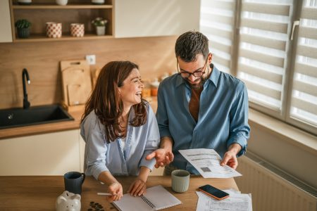 Couple sitting at home managing personal finance, reviewing bills and documents, planning expenses, and saving money in a piggy bank on the kitchen table