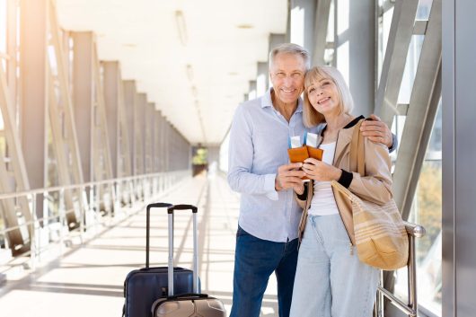 Cheerful mature couple embracing in bright airport corridor, holding passports with boarding passes, happy spouses smiling warmly with luggage in background
